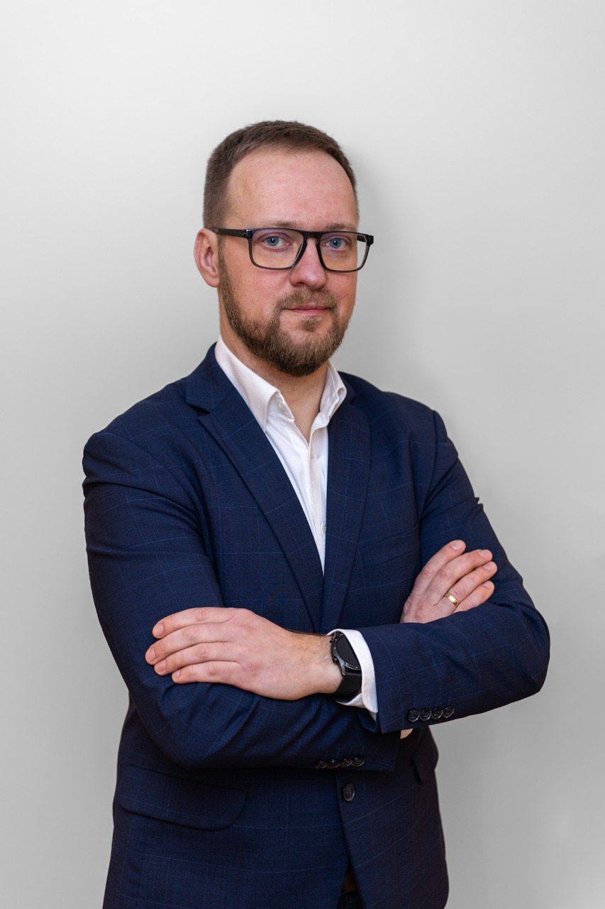 Portret mężczyzny w średnim wieku, w okularach i ciemnogranatowym garniturze z białą koszulą. Mężczyzna stoi na jasnym tle ze skrzyżowanymi rękami na piersi i uśmiecha się lekko w stronę aparatu.